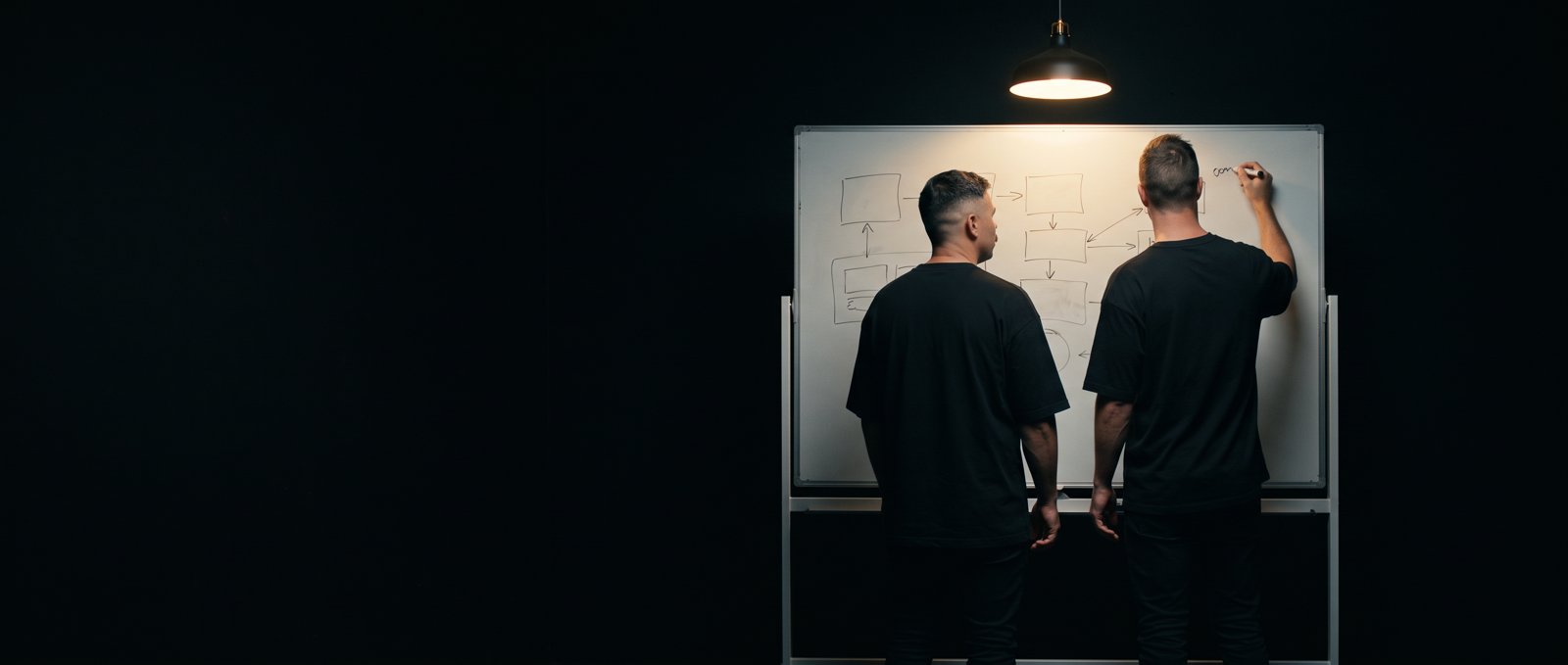 Two men in black t-shirts, seen from behind, sketching a flow diagram on a whiteboard under a single hanging lamp in a dark room.