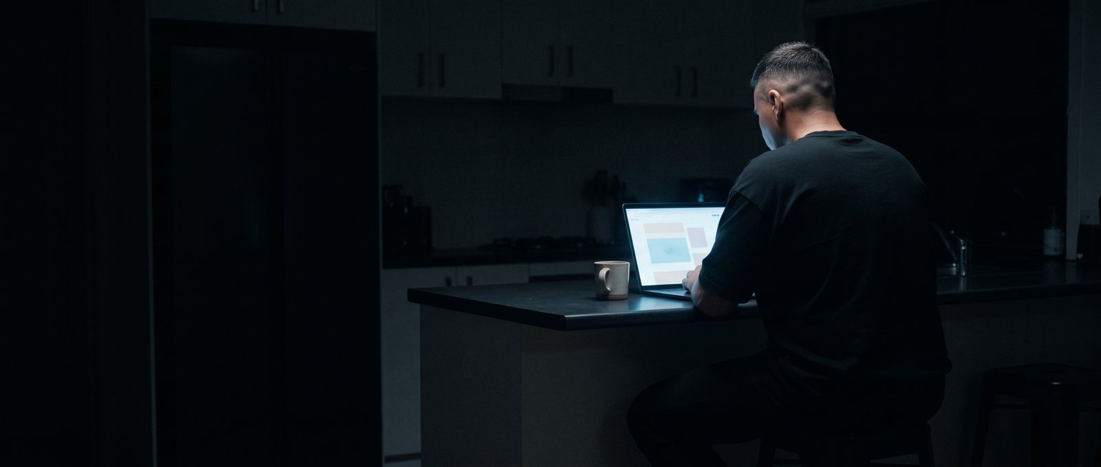 A man in a black t-shirt, seen from behind, works on a laptop at a kitchen bench in a dark room, a coffee cup beside him.